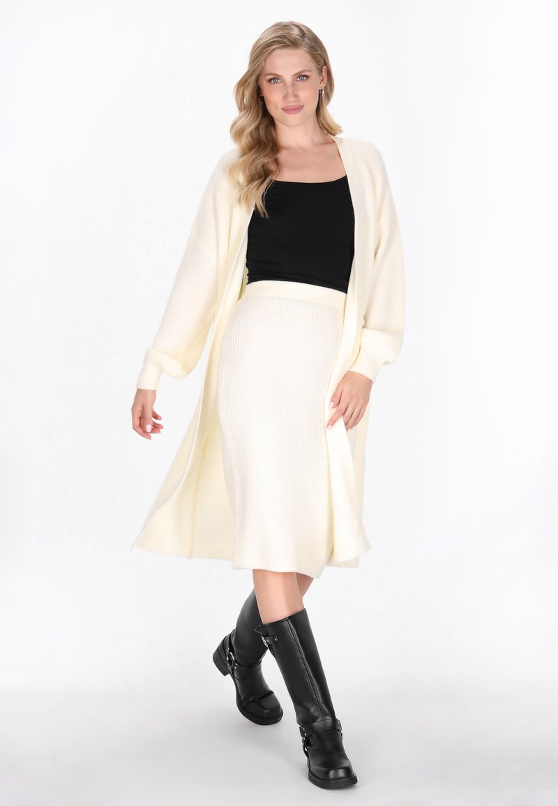 Langer, gerippter Cardigan in Cremeweiß, kombiniert mit einem passenden gerippten Rock. Schwarzes enges Oberteil und knielange schwarze Lederstiefel.