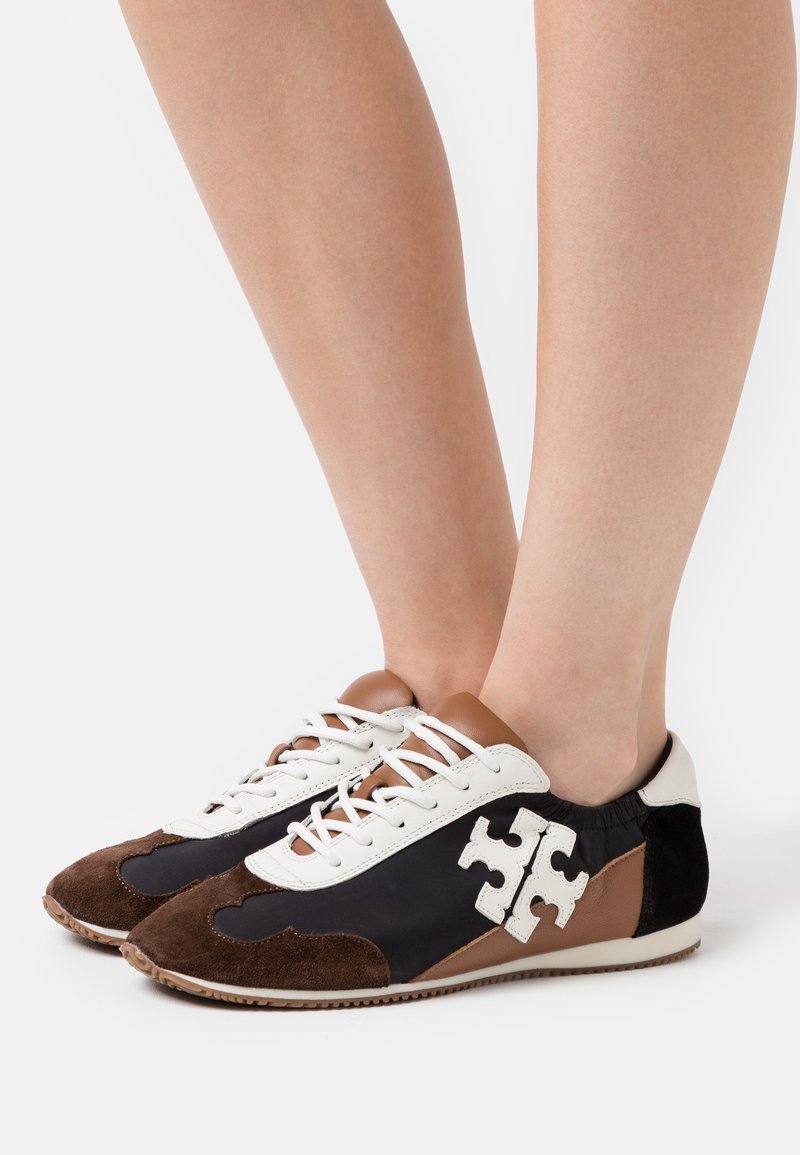 Pieds portant des baskets basses marron, noires et blanches avec des lacets à l'avant et un logo blanc en forme de pièce de puzzle sur le côté.
