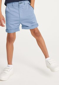 Hellblaue Shorts in maßgeschneidertem Design, mit umgeschlagenen Säumen und Seitentaschen mit kontrastfarbener Einfassung, kombiniert mit weißen Sneakers.