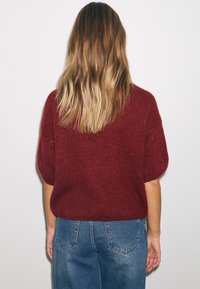 Naine, kes pöörab selja, pikkade ombre juustega, punast kudumit lühikeste varrukatega kampsunit ja siniseid teksapükse kandmas, valgel tavalise tausta ees.