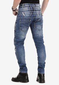 Blaue Jeans mit schmaler Passform, beschädigtem Finish, mehreren Taschen und Kontrastnähten. Verfügt über einen mittelhohen taillierten Bund und schmal zulaufende Beine.