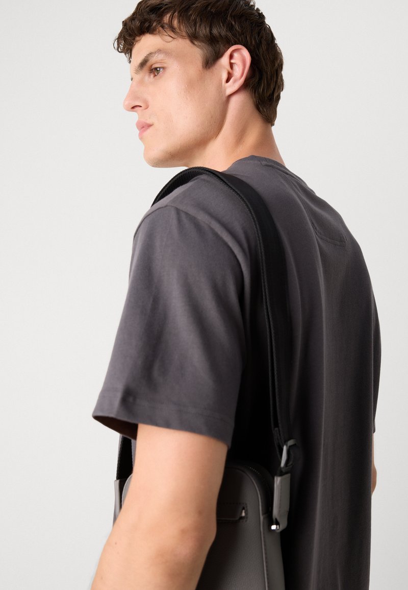 T-shirt gris et sac gris avec une sangle noire. Le sac présente une texture lisse et une forme épurée, positionné contre un fond neutre.