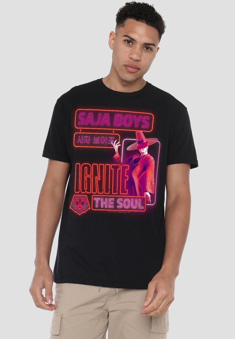 Young man wearing black t-shirt with neon-style text "SAJA BOYS," Korean script, "IGNITE THE SOUL," and a figure in a large hat in red tones.
