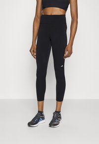 Črne športne leggins hlače z visokim pasom, raztegljivim blagom in žepom ob strani. Ujemajo se s črnimi in modrimi tekaškimi čevlji.