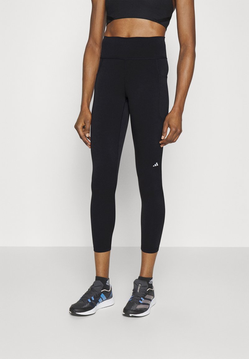 Črne športne leggins hlače z visokim pasom, raztegljivim blagom in žepom ob strani. Ujemajo se s črnimi in modrimi tekaškimi čevlji.