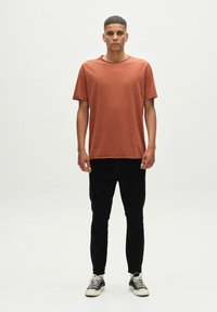 Rövid ujjú, rozsdaszínű t-shirt, laza fazonban, fekete, slim fit nadrággal és fekete-fehér sportcipővel párosítva. Egyszerű dizájn, minta nélkül.