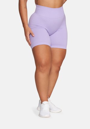 Kobieta w wysokich lawendowych szortach rowerowych i białych trampkach, stojąca z jedną ręką opartą na udzie na jednolitym tle.