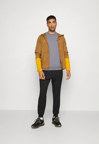 Brun lättviktsjacka med huva och gula ärmar över en grå tröja, kombinerad med svarta mjukisbyxor och svarta sportskor.