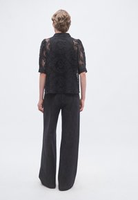 Top in pizzo nero con maniche a sbuffo, caratterizzato da motivi floreali, abbinato a pantaloni scuri ampi in tessuto a trama. Vista posteriore in mostra.