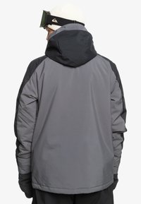 Veste isolante grise avec une capuche et des accents noirs. Elle possède une texture lisse et une coupe standard, conçue pour les activités par temps froid.