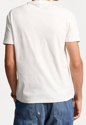 Mężczyzna w zwykłej białej koszulce z krótkim rękawem i niebieskich dżinsach, pokazany z tyłu, stojący z rękami w kieszeniach.