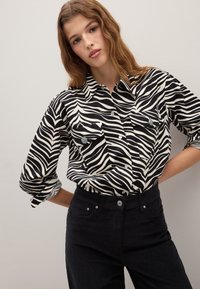 Camisa de estampado de cebra en blanco y negro con diseño de cuello, dos bolsillos en el pecho, mangas enrolladas y detalles de botones, combinada con pantalones oscuros.