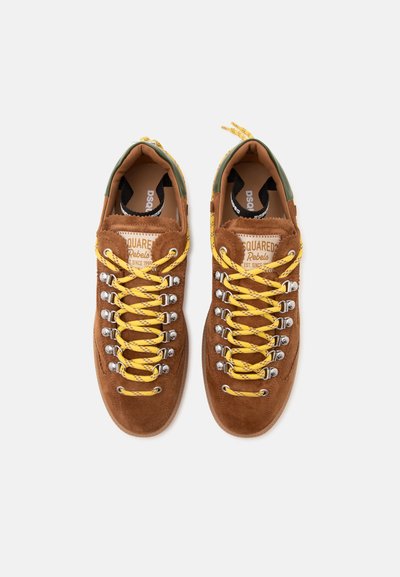 Ζευγάρι καφέ sneakers από σουέντ με κίτρινα κορδόνια, μεταλλικά κρικάκια και πράσινες λεπτομέρειες στη φτέρνα, σε θέαση από πάνω σε λευκό φόντο.