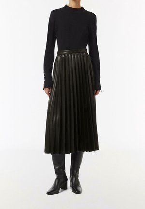 Mujer con top negro de manga larga, falda midi plisada de cuero negro y botas negras altas con tacón, de pie sobre un fondo blanco.