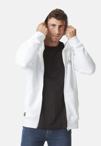 Haut à capuche blanc zippé avec une poche avant, capuche ajustable et poignets côtelés, porté sur un t-shirt noir, associé à un jean bleu.