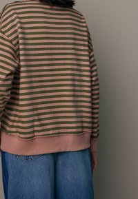Persona che indossa un maglione a righe orizzontali verdi e rosa e jeans blu larghi, in piedi contro uno sfondo grigio chiaro uniforme.