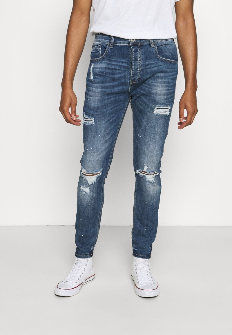 Homme portant un jean skinny bleu délavé et des baskets montantes blanches, debout devant un fond blanc uni.