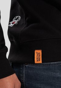 Sudadera negra con un personaje bordado blanco en el costado y una etiqueta naranja "WRONG FRIENDS" en el dobladillo. Puños y dobladillo de canalé.