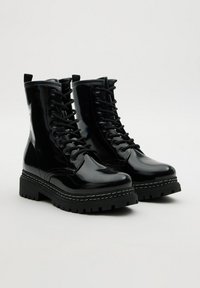 Botas de combate negras brillantes con cordones y suelas gruesas, con costuras blancas en la entresuela, mostradas sobre un fondo claro y liso.