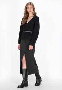 Zwart, fluweelachtig cardigan met een V-hals, met een ceintuur op de taille, gecombineerd met een zwarte denim rok met een voorknipsel en hoge zwarte laarzen tot de knie.