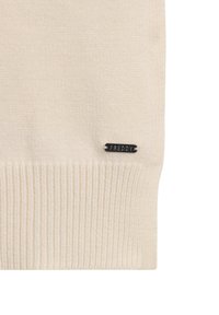Maglione color crema con texture a coste, caratterizzato da un'etichetta metallica nera incisa con la scritta "FREDDY" sul lato inferiore sinistro.