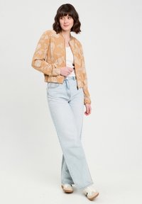 Tan bomber jacket dengan pola bunga putih, dengan ritsleting penuh, dan manset berserat; dipadukan dengan jeans tinggi biru muda dan sepatu sneaker beige.