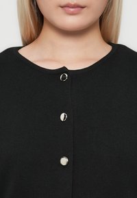 Close-up of a person wearing a black buttoned sweater with three shiny round silver buttons and a round neckline.