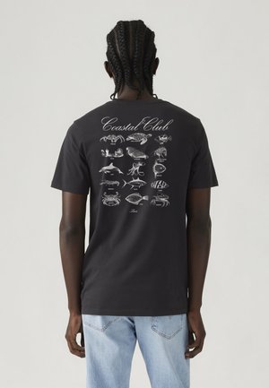 Asmuo su pintais plaukais, dėvintis juodą marškinėlį, rodantis atspausdintą nugaros pusę su įvairiais pažymėtais jūros gyvūnais ir užrašu „Coastal Club“.