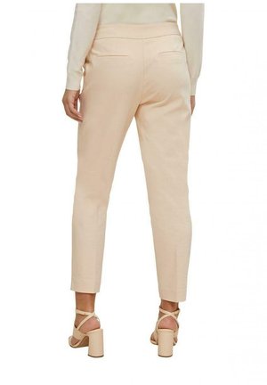 Žena oblečená v béžových úzkých kalhotách po kotníky a odpovídajících sandálech na podpatku, zobrazena zezadu na bílém pozadí.