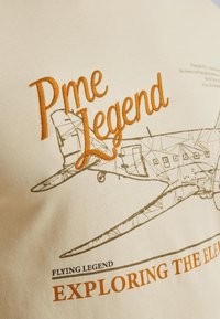 Kremowa bawełniana koszulka z haftowanym napisem "Pme Legend" w kolorze pomarańczowym oraz szczegółową grafiką samolotu w kolorze brązowym, z napisem "Flying Legend" poniżej.