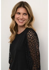 Haut noir avec des manches en dentelle présentant un motif floral. Le corps est uni, tandis que les manches sont semi-transparentes avec des détails complexes.