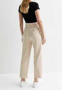 Vrouw met lang blond haar draagt hoogwaardige beige broek, zwart T-shirt met korte mouwen en witte sneakers, staand tegen een witte achtergrond.