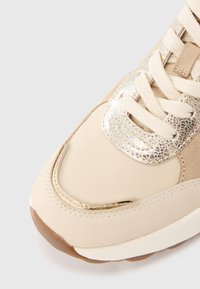 Tenisica ima gornji dio od bež tkanine s detaljima od brušene kože, metalik zlatnim ukrasima i krem vezicama; zaobljeni vrh i gumeni potplat.