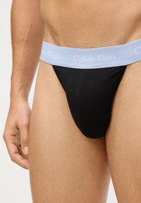 Crni tkaninski tanga gaćice s svijetloplavim elastičnim pojasom na kojem je u bijelom napisan logo 'Calvin Klein'. Glatka tekstura, kroj koji se prilagođava tijelu.