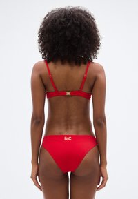 Κόκκινο μπικίνι με λεπτές ρυθμιζόμενες τιράντες και χρυσή λογότυπη λεπτομέρεια. Λείο, ελαστικό ύφασμα με μίνιμαλ σχεδιασμό και μέτρια κάλυψη.