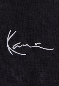 Texte en cursive brodé en blanc sur un tissu noir affichant le mot « Kani » avec un « K » stylisé de manière distinctive.
