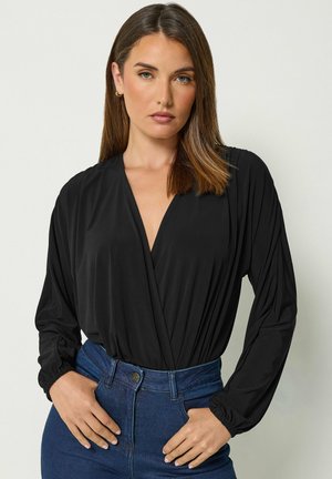 Frau mit glatten braunen Haaren, die ein schwarzes langärmliges V-Ausschnitt-Top trägt, in hoch taillierte blaue Jeans gesteckt, steht mit Händen in der Hüfte.