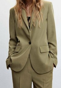 Blazer verde oliva su misura con chiusura a un solo bottone, rever a punte e due tasche anteriori. Tessuto liscio e vestibilità strutturata.