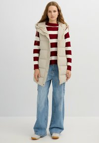 Chaleco acolchado beige con capucha, cremallera frontal y botones a presión, usado sobre un suéter de rayas rojas y blancas y jeans azules holgados.