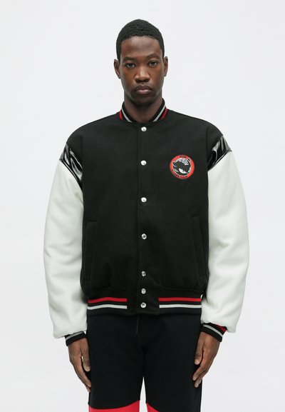 Color-blocked varsity jacket in black and white, featuring a ribbed collar, snap buttons, and a round patch on the chest. Textured sleeves with accents.
