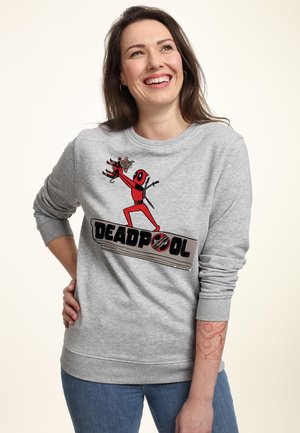 Kvinde smilende iført en grå sweatshirt med Deadpool-grafik, holder en lille figur og blå jeans, står mod en ensfarvet baggrund.