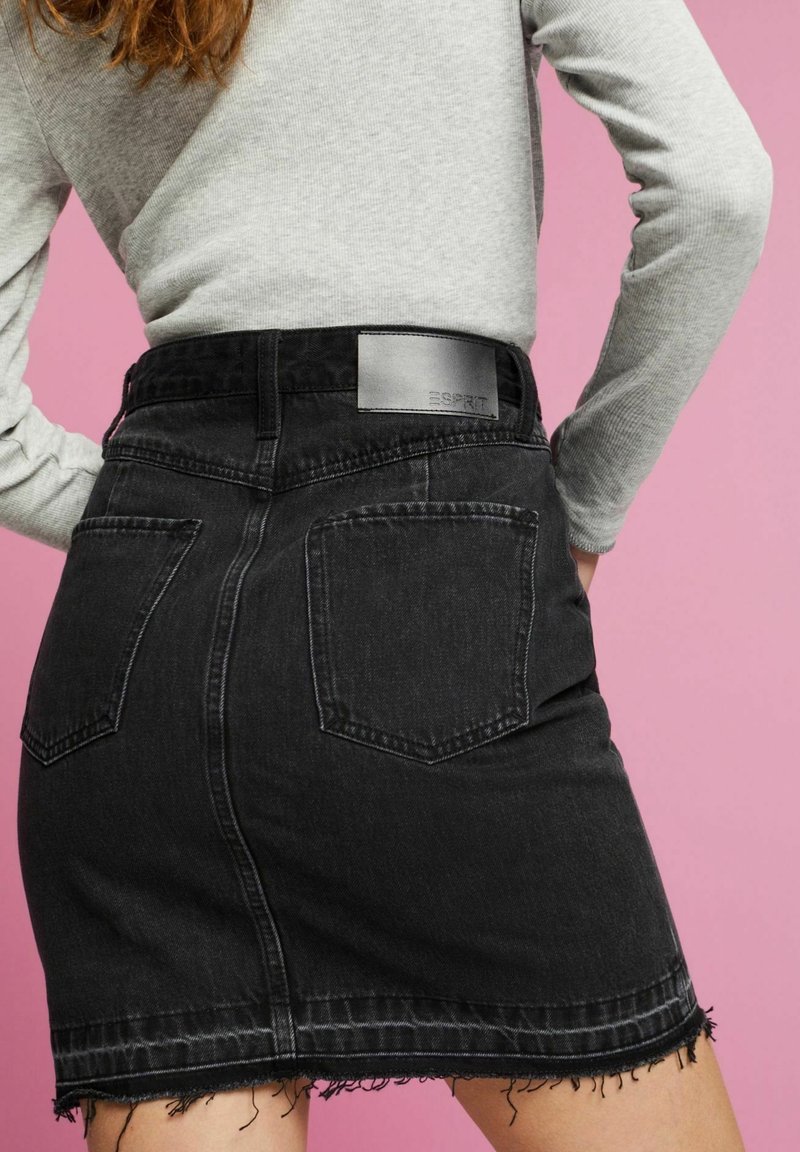 Personne portant un haut gris à manches longues et une jupe en denim noire avec des poches arrière et un ourlet effiloché, sur un fond rose.