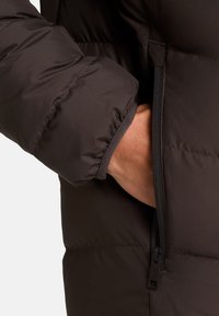 Giacca puffa marrone con tasca con zip in vita. Il materiale è liscio e isolato, con polsini arricciati per garantire calore.