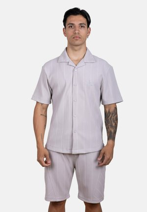 Hombre con cabello corto y negro que lleva una camisa de manga corta gris claro abotonada y pantalones cortos a juego, de pie contra un fondo liso.