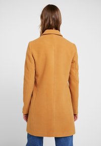 Manteau couleur moutarde avec un col structuré et une texture lisse. Présente une silhouette cintrée et est mi-longueur, avec un design épuré et minimaliste.