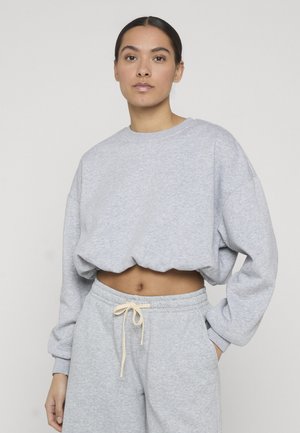 Fiatal nő világosszürke, rövidített pulóvert és hozzá illő melegítőnadrágot visel bézs színű húzózsinórral, egyszínű világos háttér előtt áll.