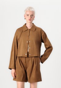 Bruine cropped jacket met een kraag, zilveren drukknoopjes en brede mouwen, gecombineerd met bijpassende geplooide shorts; gladde textuur.