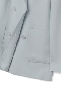 Blazer azul claro con cuatro botones redondos y dos bolsillos delanteros tipo parche, mostrado parcialmente abierto sobre un fondo blanco.