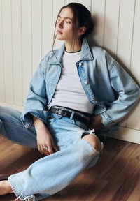 Ung kvinna som sitter på ett trägolv mot en ljus vägg, klädd i en ljus jeansjacka och trasiga jeans, ser eftertänksamt åt sidan.