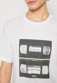 Weißes Baumwoll-T-Shirt mit einem Grafikdesign von zwei VHS-Kassetten, beschriftet mit "Meine schlechten Tage" und "Meine guten Tage" in schwarzem und blauem Text.